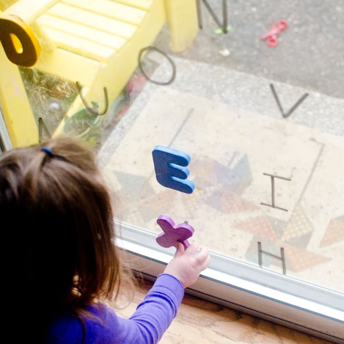 A child reaches for a purple letter X stuck to a window. There are dry erase marker letters on the glass and other foam letters stuck to it.