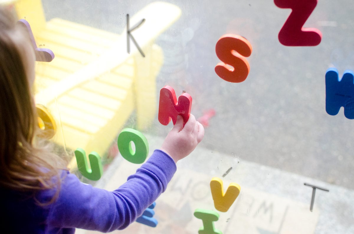 A child reaches to add a red letter N to a window. There are dry erase marker letters on the glass and other foam letters stuck to it.