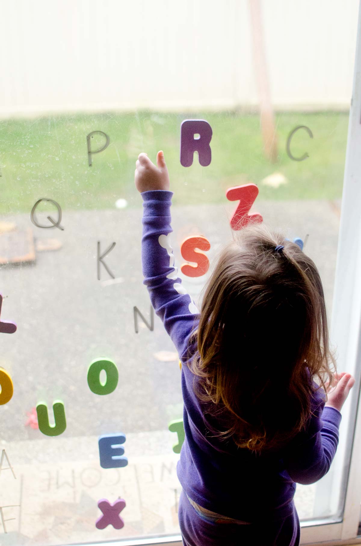 A child reaches for a purple letter R stuck to a window. There are dry erase marker letters on the glass and other foam letters stuck to it.