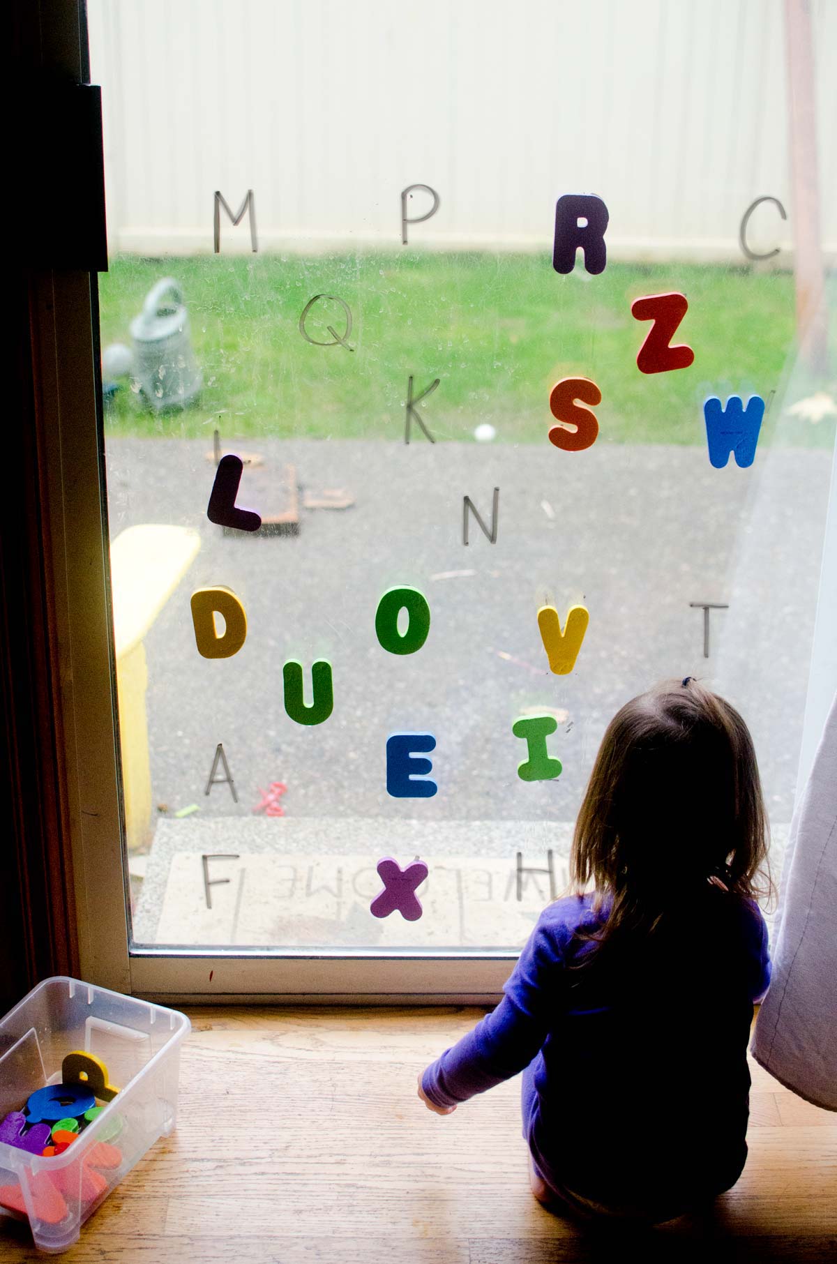 A child squats on the floor in front of a sliding glass door placing foam letters onto to. There are many letters on the glass.