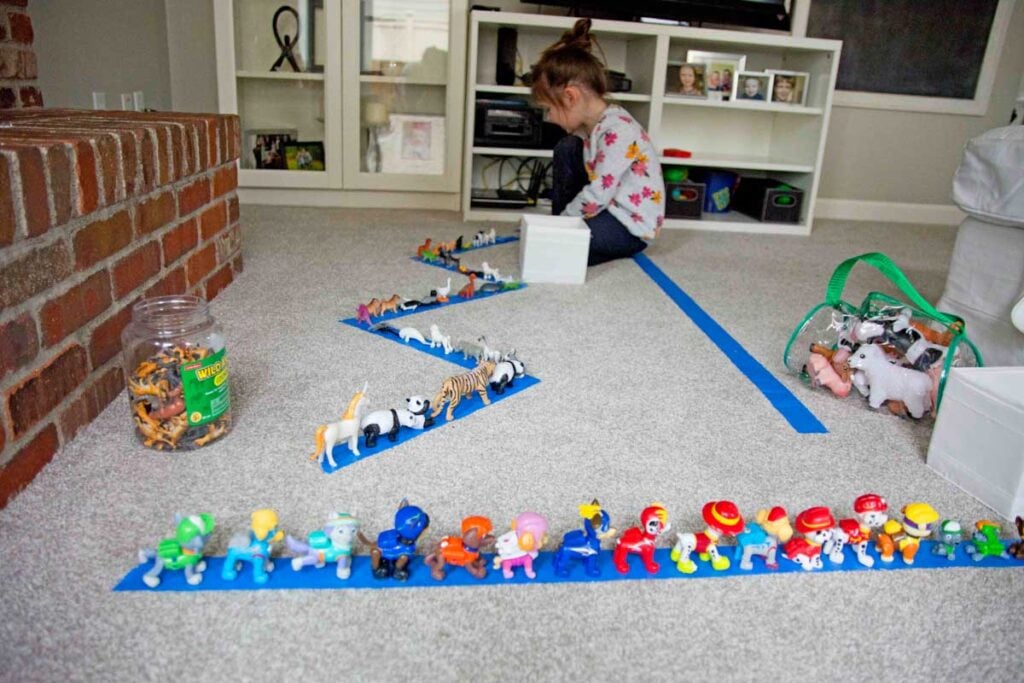 A child places toys onto a strip of blue tape. There are more animals on tape strips behind her.
