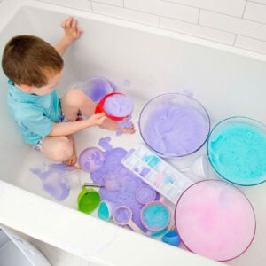 A child in a bathing suit sits in a bathtub. There are bowls of purple, blue, and pink bubble foam around him.