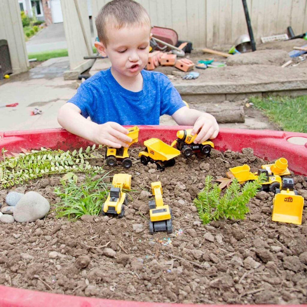 A child in a blue shirt is making a "vrooming noise" as they move small construction trucks around a sensory bin full of dirt and yard trimmings.