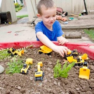 A smiling child in a blue shirt moves small construction trucks around a sensory bin full of dirt and yard trimmings.