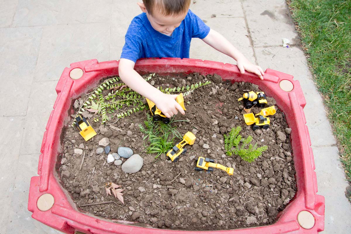 Overhead photo of child in a blue shirt moving small construction trucks around a sensory bin full of dirt and yard trimmings.
