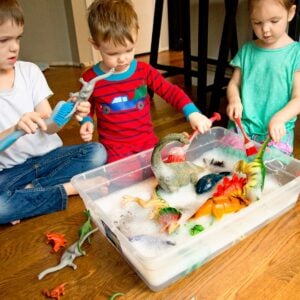 Three kids sit around a bin of water with plastic dinosaurs. They are washing the dinosaurs.