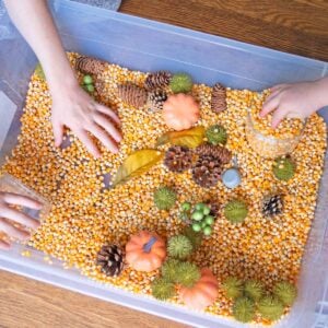Three kid hands reach in a sensory bin made from popcorn kernels and fake fall decorations.