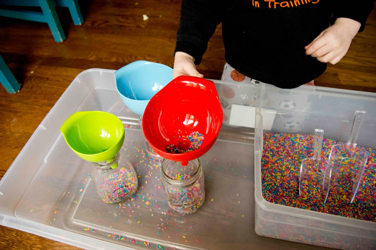 A child grabs at a red funnel while playing in a rainbow rice sensory bin.