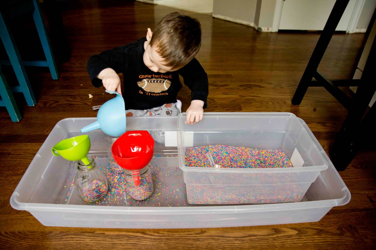 A child picks up a blue funnel and tips it. He is playing with rainbow rice in a sensory bin.