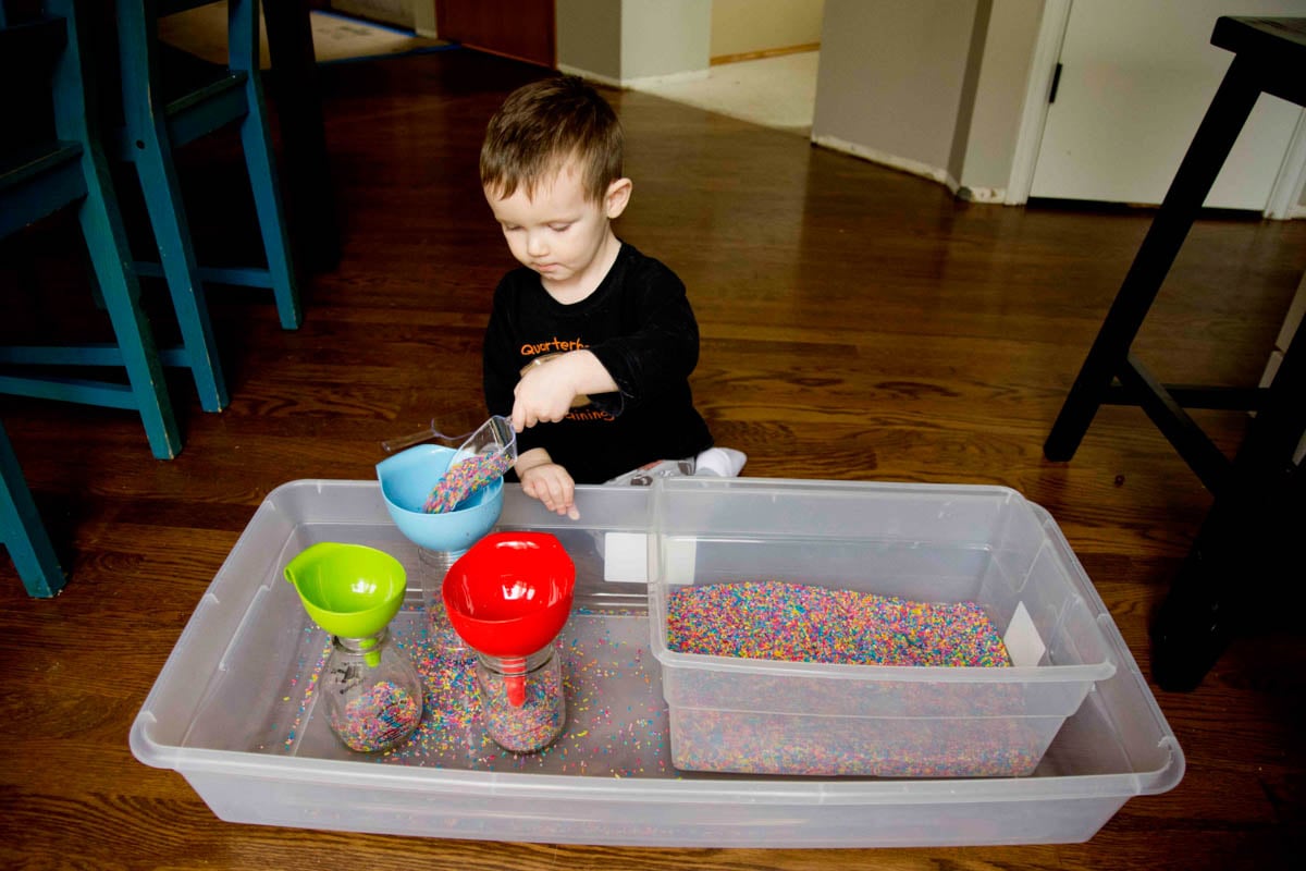 A toddler concentrates as he adds rainbow rice to a blue funnel. He is 2 and wearing pajamas.