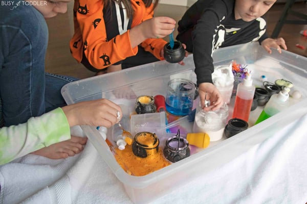 A child learns forward in a potion bin to scoop baking soda; another child has a fizzing cauldron of orange goo.