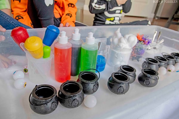 A before photo of a Halloween potion bin with mini cauldrons, colorful vinegar, shaving cream, spiders, and turkey basters.