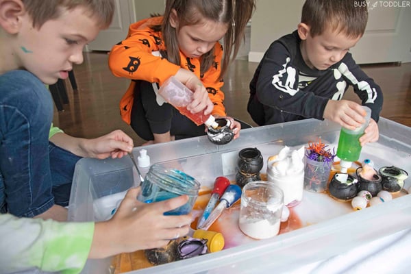 Three kids playing at a Halloween potions activity with mini cauldrons, shaving cream, baking soda, and soap visible.