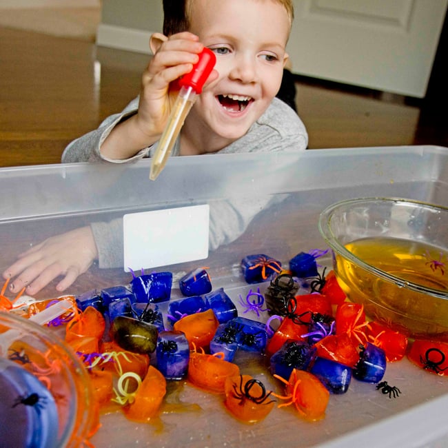 A child laughing with a small turkey baster over a collection of orange and purple ice cubes with spiders frozen inside.