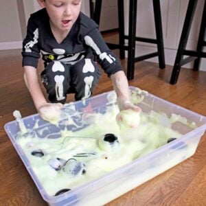 A child in skeleton pajamas playing with a Halloween themed sensory bin made of green bubble foam.