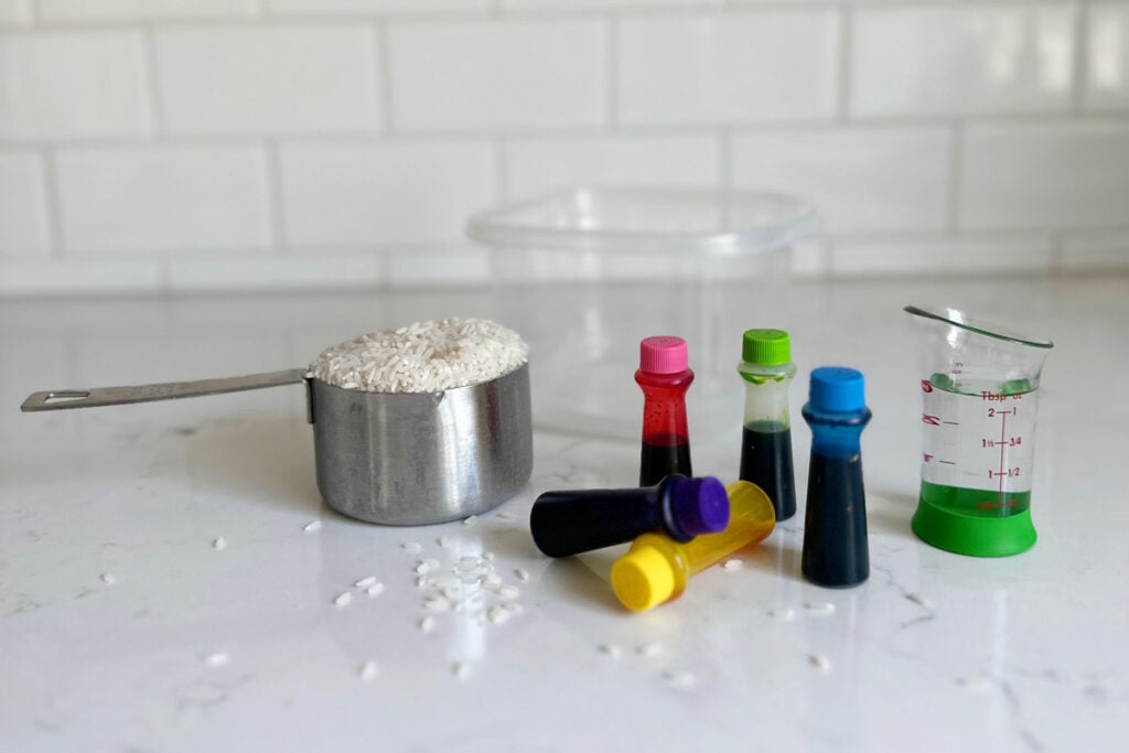 The supplies for how to dye rice are on a counter: rice, food coloring, vinegar, and a container.