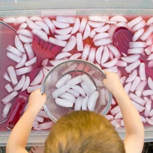 Child playing in a storage container (sensory bin) with ice cubes floating in red-dyed water. Kitchen tools and colanders are in the image.