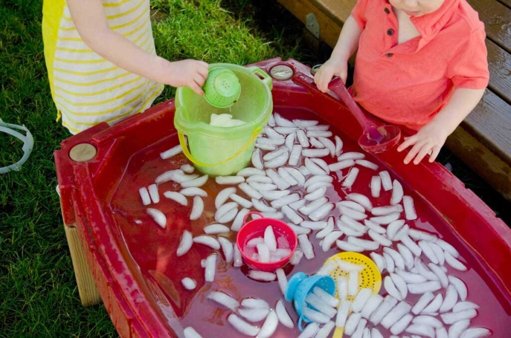 Two children play at a sand table full of ice cubes and water. One is scooping ice while the other holds a bucket.
