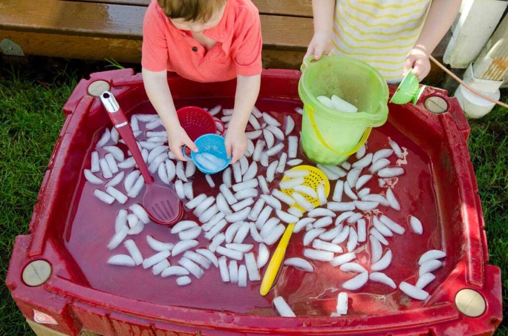 Two children play at a sand table full of ice cubes and water. One is scooping ice while the other holds a bucket.