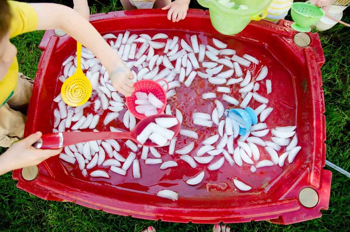 Three children play at a water table filled with ice. One is scooping ice into a colander.