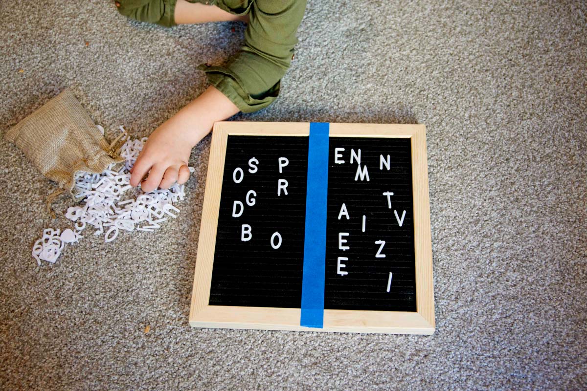 Overhead shot of a letter board with blue tape down the middle. A child's hand is placing letters onto the board into sorted groups.