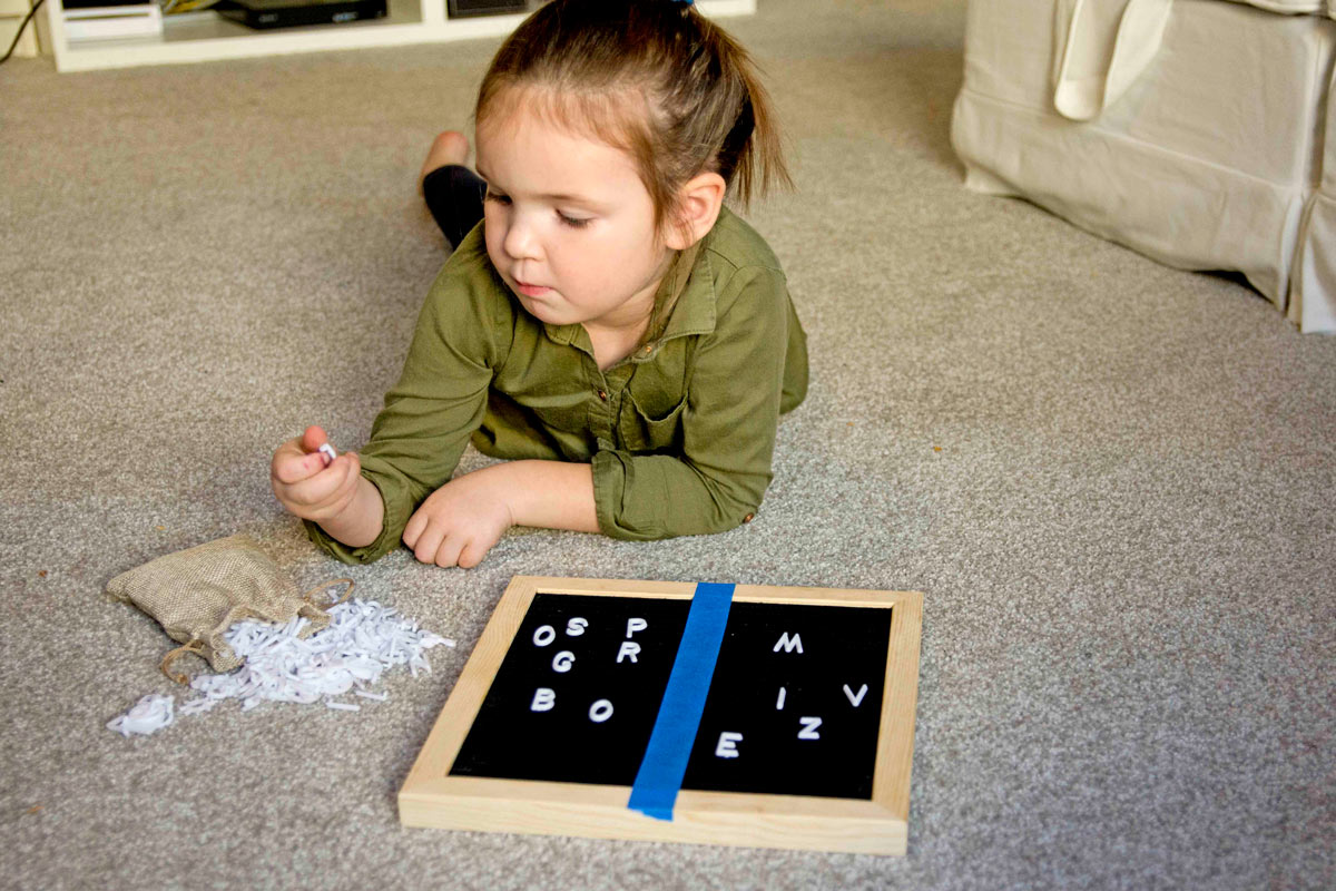 A child examines a plastic white letter to decide where to place it on a letter board. The letter board is split into two halves using blue tape.