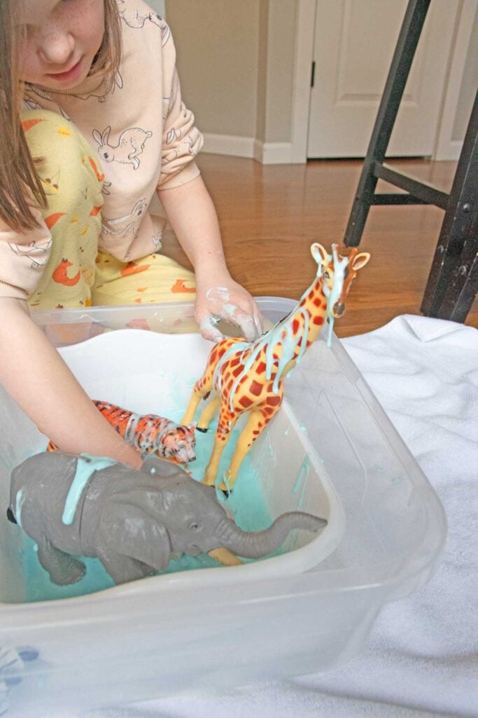 A child plays with animals in a sensory bin full of oobleck. The child is decorating a large giraffe toy.
