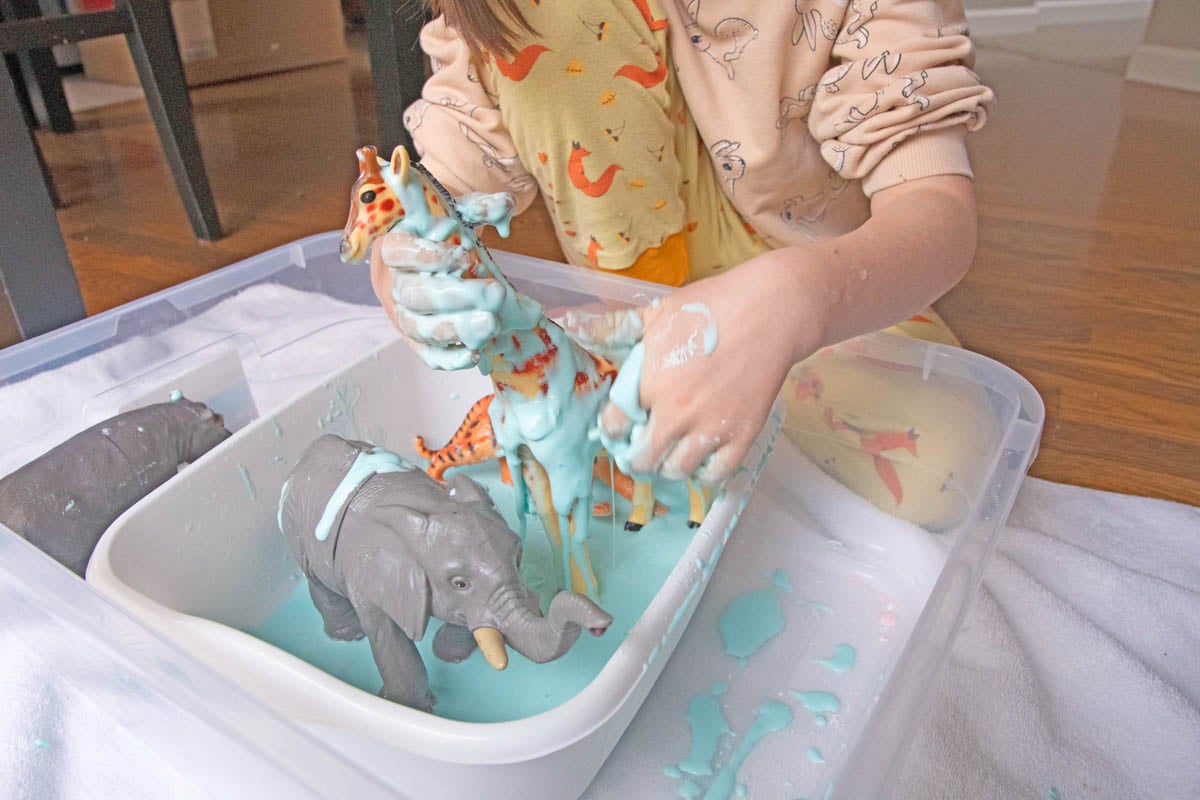 A child with hands covered in oobleck "decorate" a toy giraffe in a sensory bin.