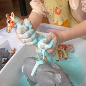 A child plays with blue oobleck in a sensory bin. There are also plastic animals in the oobleck.