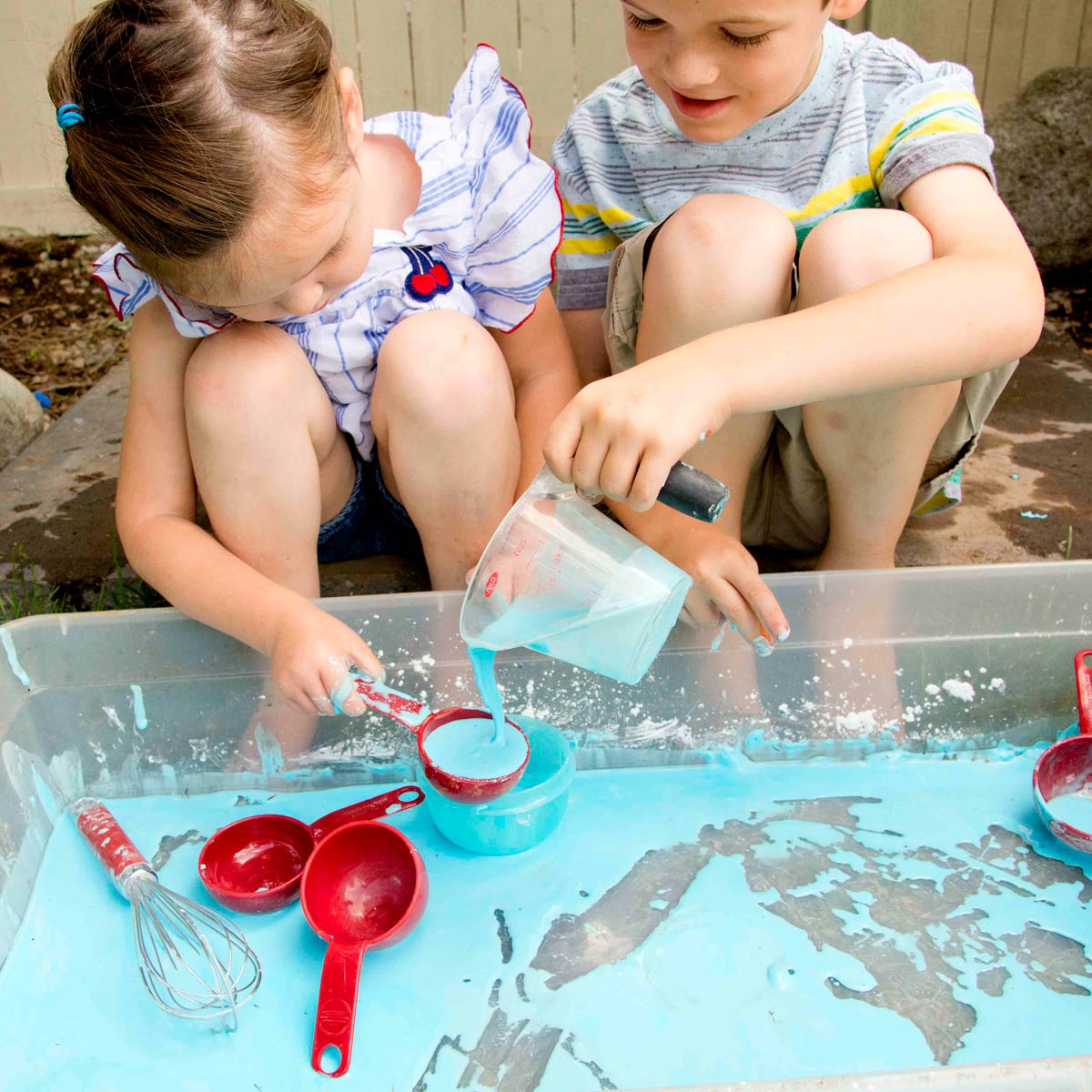 Two kids play with blue oobleck in a sensory bin. The boy is smiling as he pours oobleck into a measuring cup that the girl is holding.