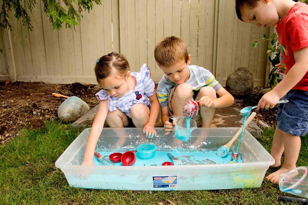 Three children play at an oobleck sensory bin outside with baking supplies.