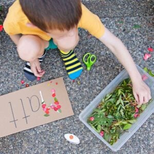 A child sits outside and reaches into a bin of yard trimmings. In front of them is a paper with their name (Matt) written on it, glue, and scissors. The child is making a collage of their name.
