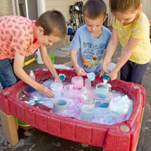 Three kids stand over a water table full science experiments bubbling in containers.