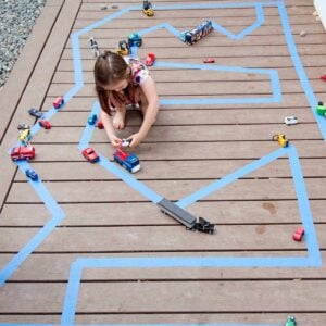 A girl plays on a deck with a blue tape road. She is driving toy cars.