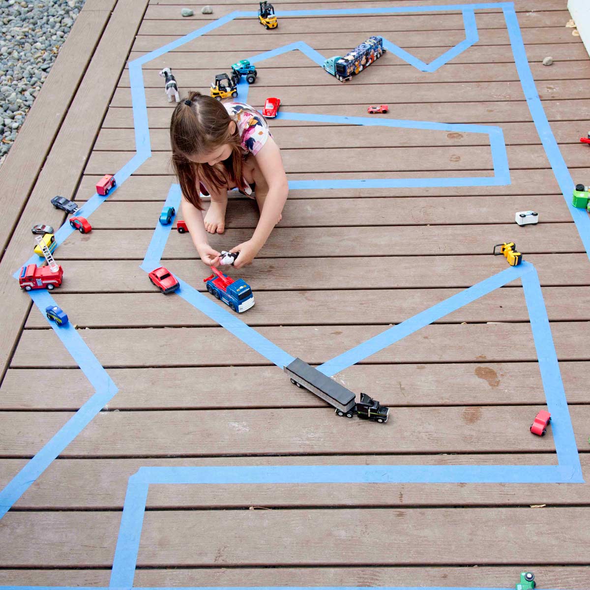 A child plays on an outdoor road made from painter's tape. There are small cars surrounding her.