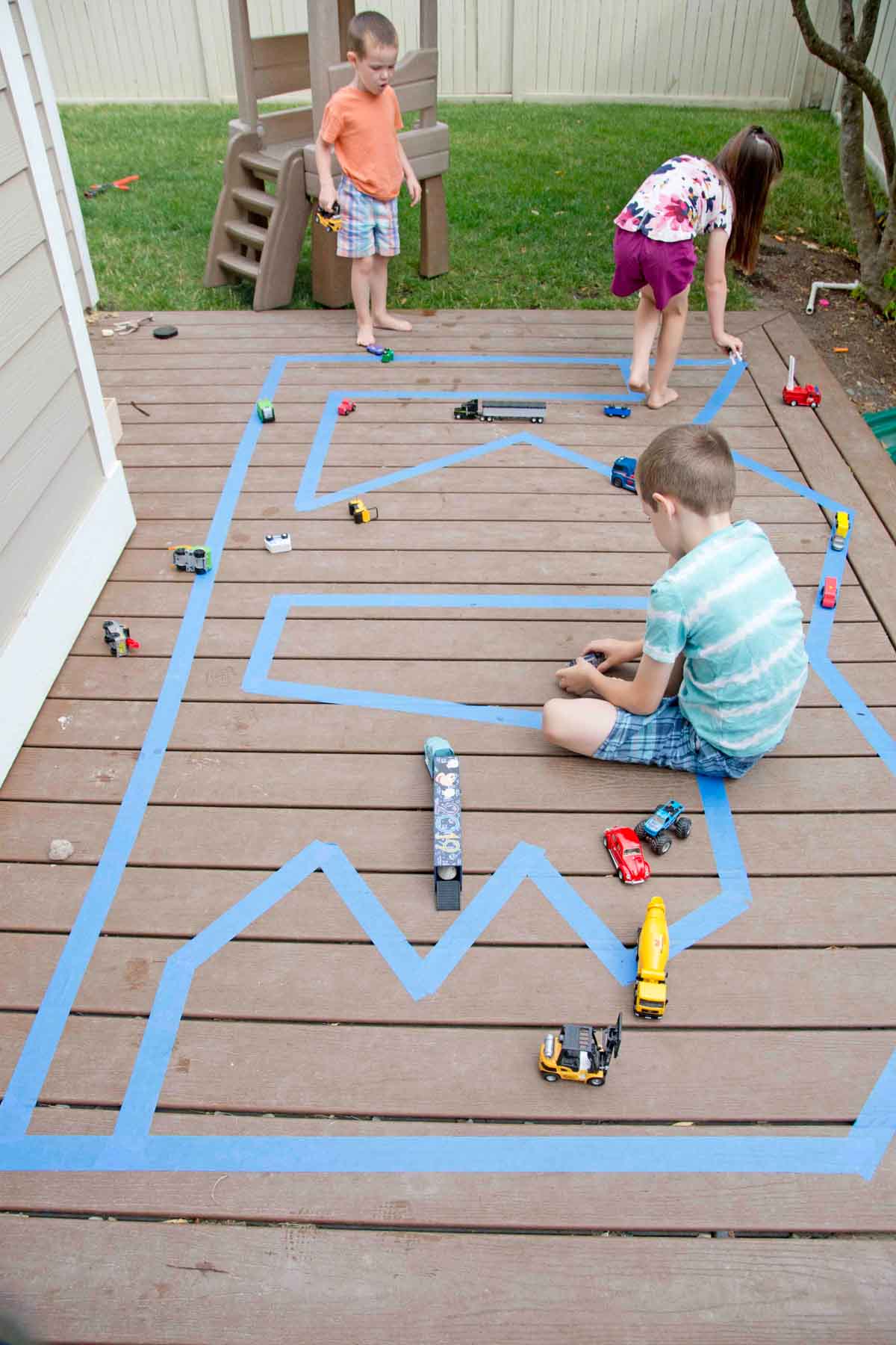 Three kids play on a deck with an outdoor road made from painter's tape. There are small cars around them.
