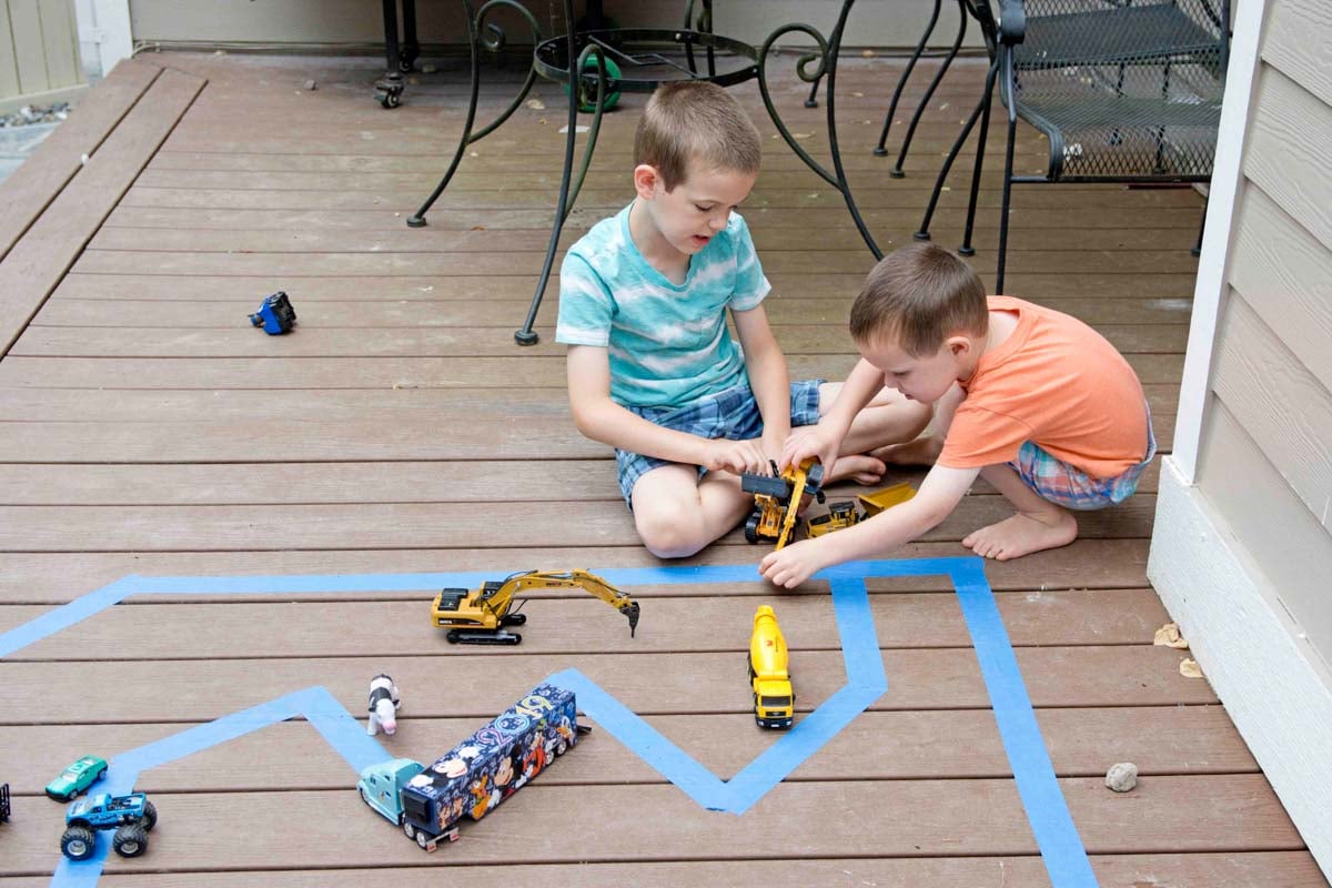 Two kids play on a back deck with a tape road. They are driving cars on the tape.