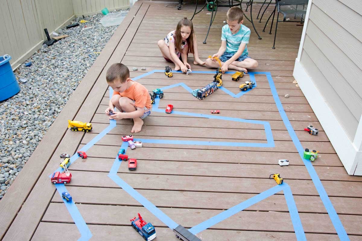Three children plays on an outdoor road made from painter's tape. There are small cars surrounding them.