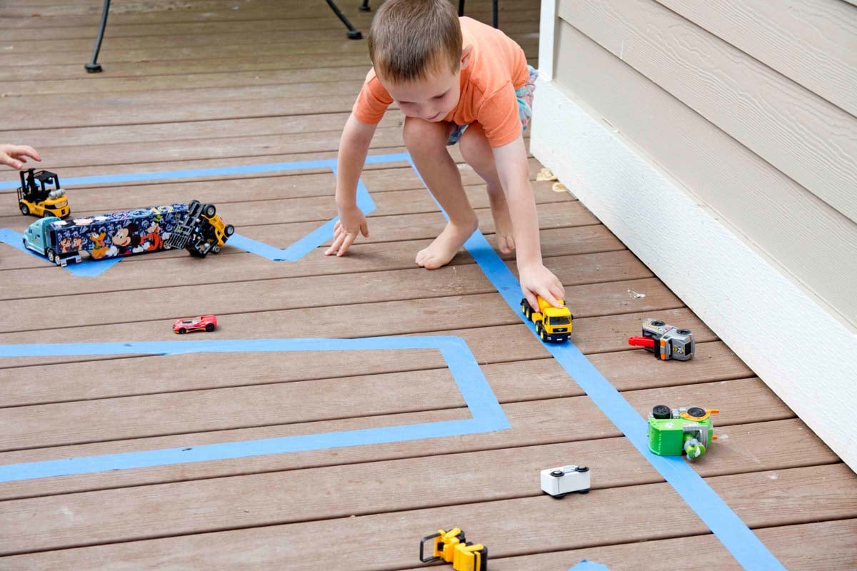 A child holds a yellow truck and pushes it along a tape road made on his deck.