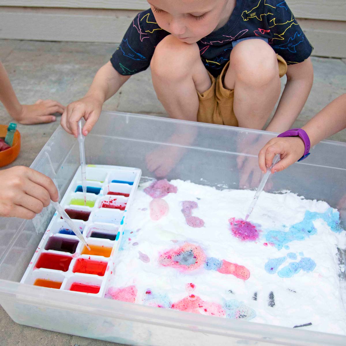 A child crouches down behind a plastic bin filled with baking soda. An ice cube tray next to him is full of colorful baking soda.