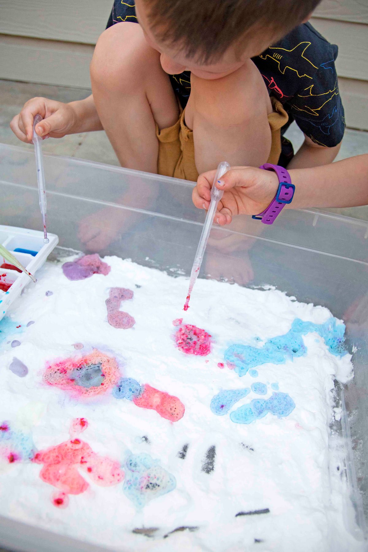 A child's hand holes a pipette and squeezes pink vinegar into a bin of baking soda.