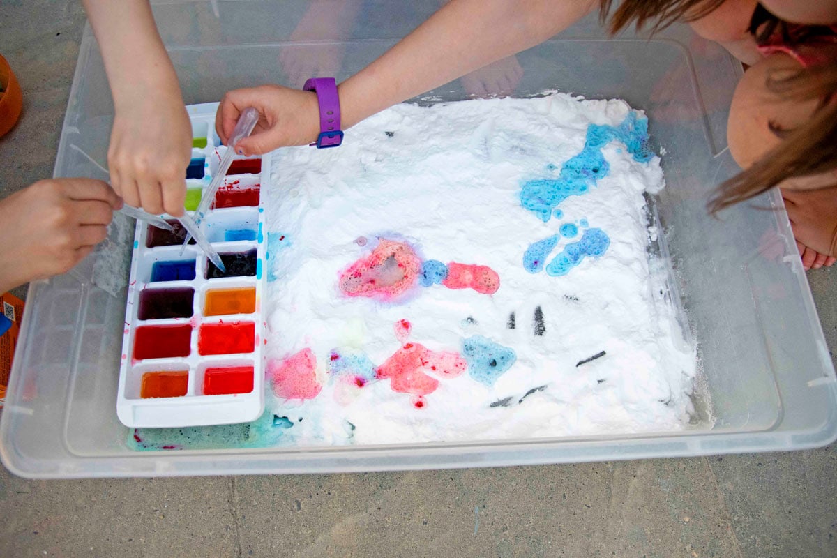 Overhead photo of a storage bin filled with baking soda. Three kid hands reach in to fill pipettes with colorful vinegar.