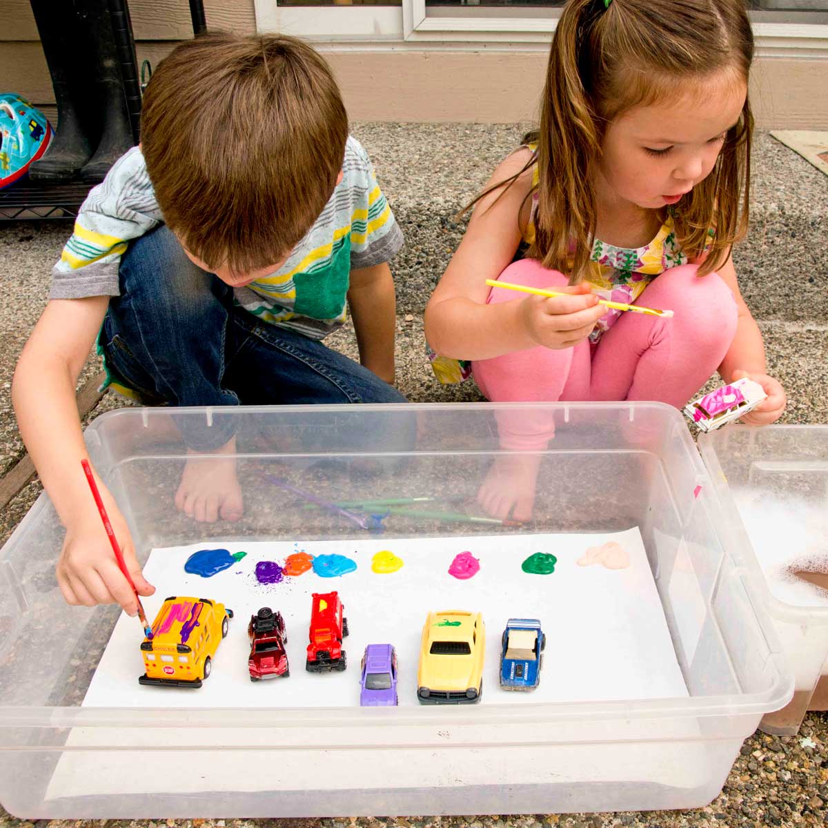 Two children sit outside holding paint brushes and metal cars. Many cars are sitting in a storage container. The children are painting the cars.