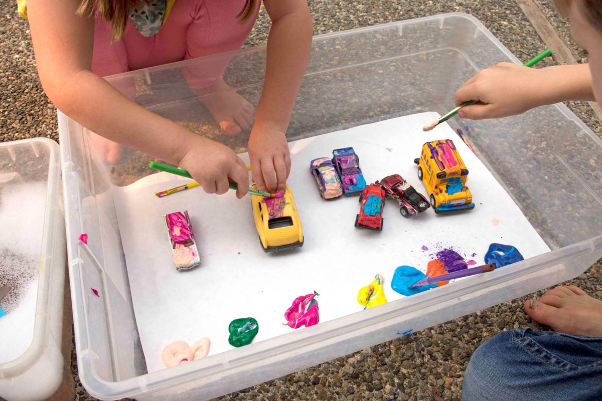 A storage container with a bunch of hot wheels cars lined up. Paint is also in the bin and children are painting the cars.
