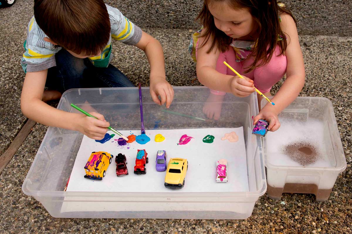 Two children sit outside holding paint brushes and metal cars. Many cars are sitting in a storage container. The children are painting the cars.