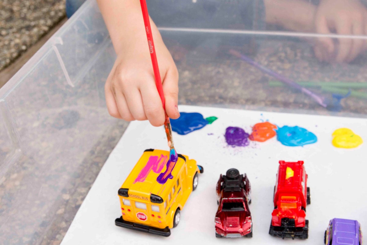 A child is swiping paint onto a yellow school bus toy. Other cars are lined up next to the school bus toy.