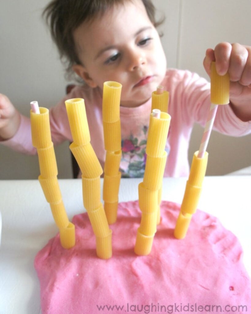 A child adds pasta to sticks in play dough.