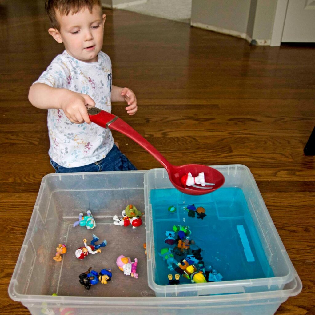 A child holds a red slotted spoon and is scooping toys from blue water into a safe space.