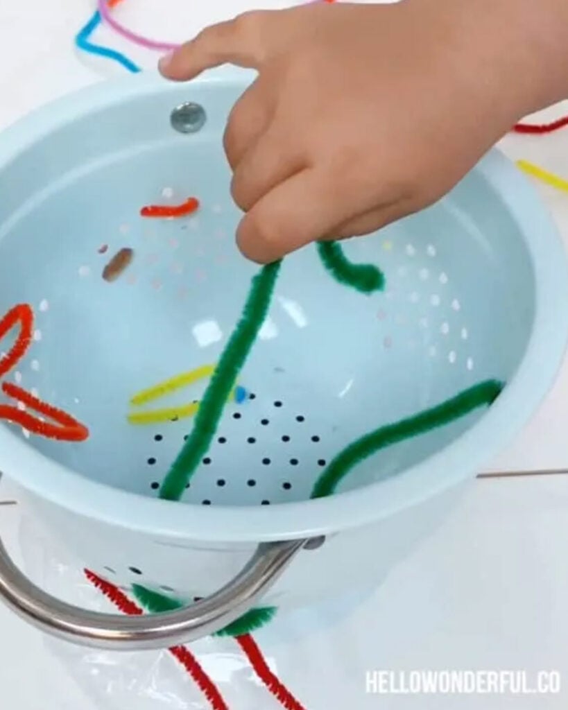 A child's hand works a pipe cleaner into a colander. 