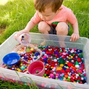 A child pours pom pom balls back into a sensory bin that is full of water.